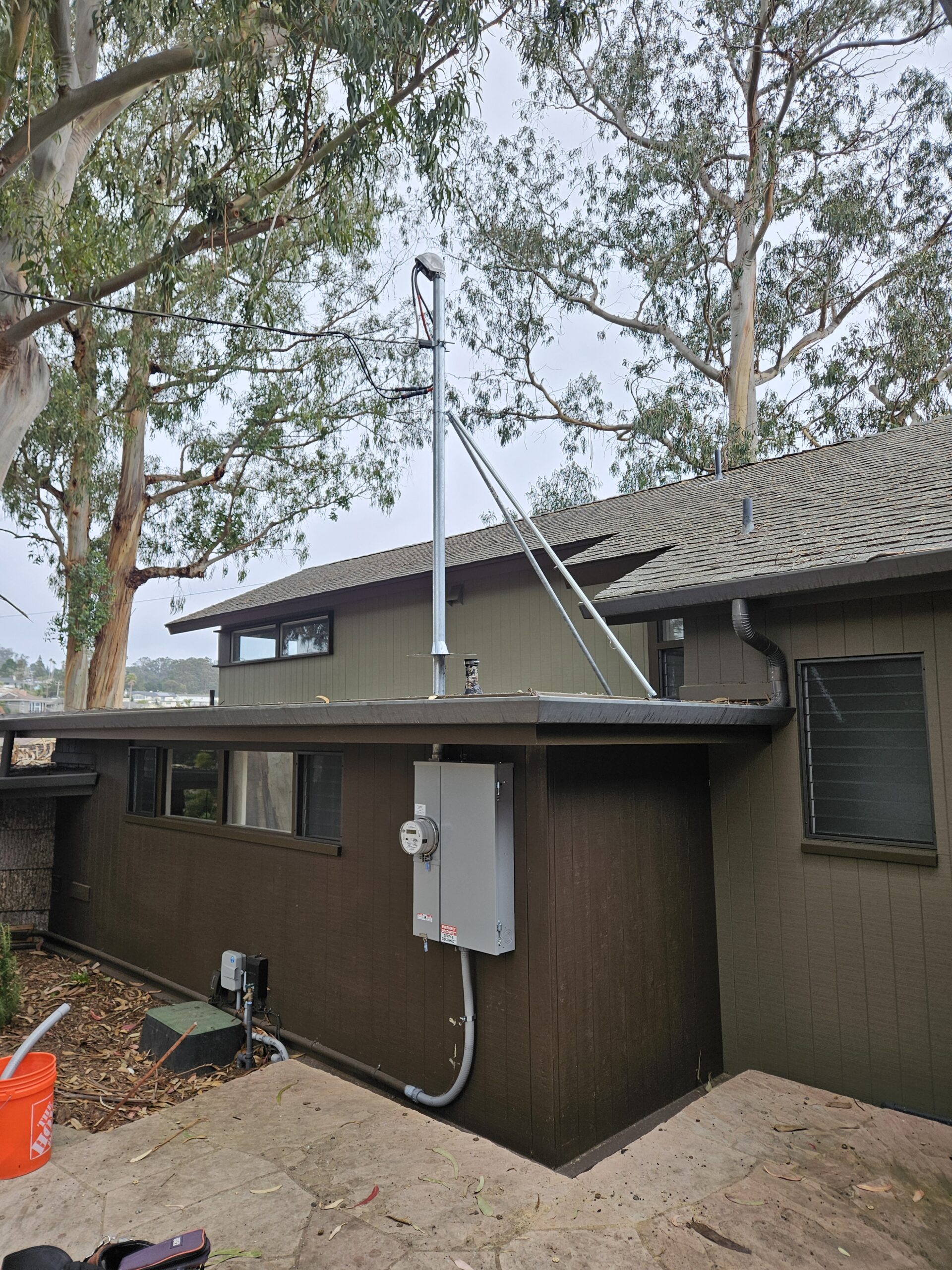 A metal pole with guy wires is mounted on the roof of a brown house, beside an electrical panel and conduit attached to the wall. Trees are visible in the background.